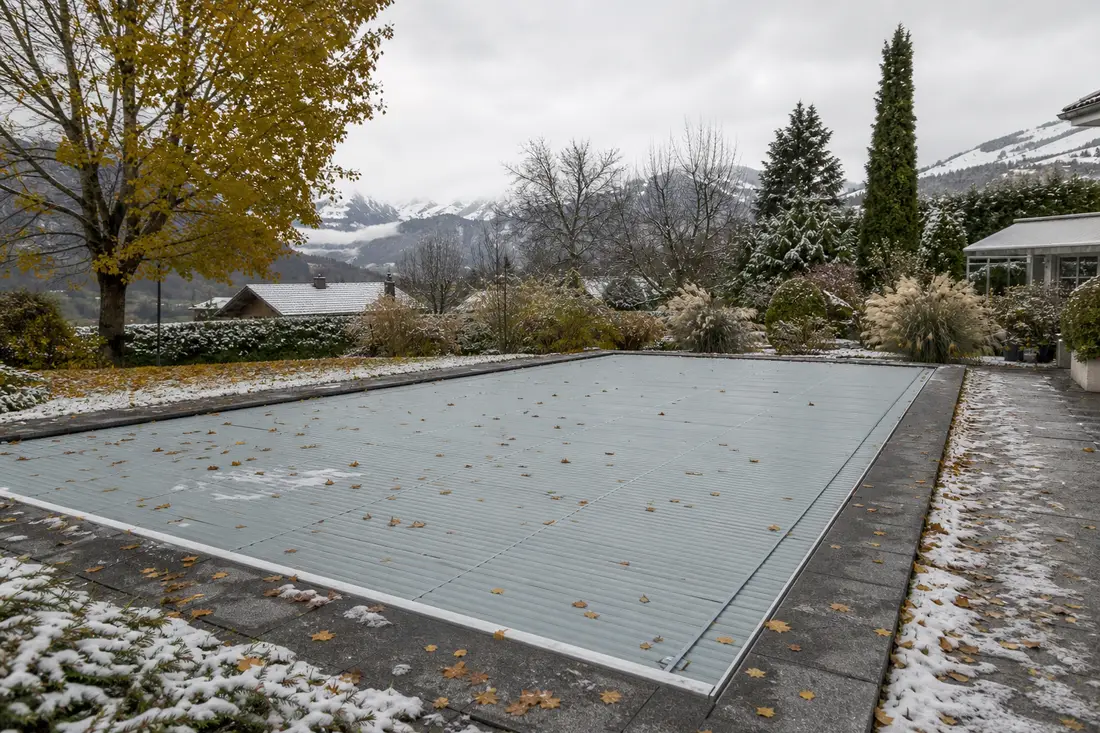 Mit einer Lamellen-Winterabdeckung geschlossener Gartenpool in verschneiter Schweizer Berglandschaft mit Herbstlaub